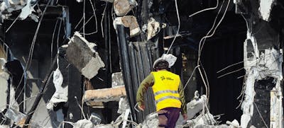 A worker in a bright yellow vest inspects a collapsed building.