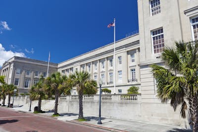 A light-colored federal building, showcasing multi-pane windows and a raised entryway adorned with decorative concrete bollards, stands just beyond a red brick road bordered by palm trees. In the center of the building's outdoor promenade, an American flag stands taller than the building, waving in the breeze.