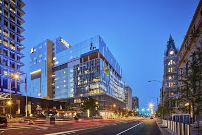 Above a bustling night scene of a roadway illuminated by streaks of vehicular traffic lights, a brightly lit multi-story building with a mirrored facade stands prominently illuminated.