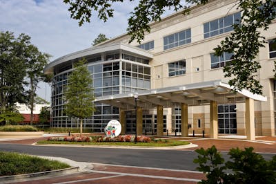 Front view of a multi-story building featuring a prominent glass façade spanning from floor to roof, with a shaded walkway leading to the entrance. The circular driveway is adorned with decorative colored pavers, bordered by well-maintained grass and flower beds.