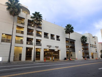 The exterior of a multilevel, well-lit and light beige colored parking garage. Tall palm trees stand out front, adding to the aesthetic.